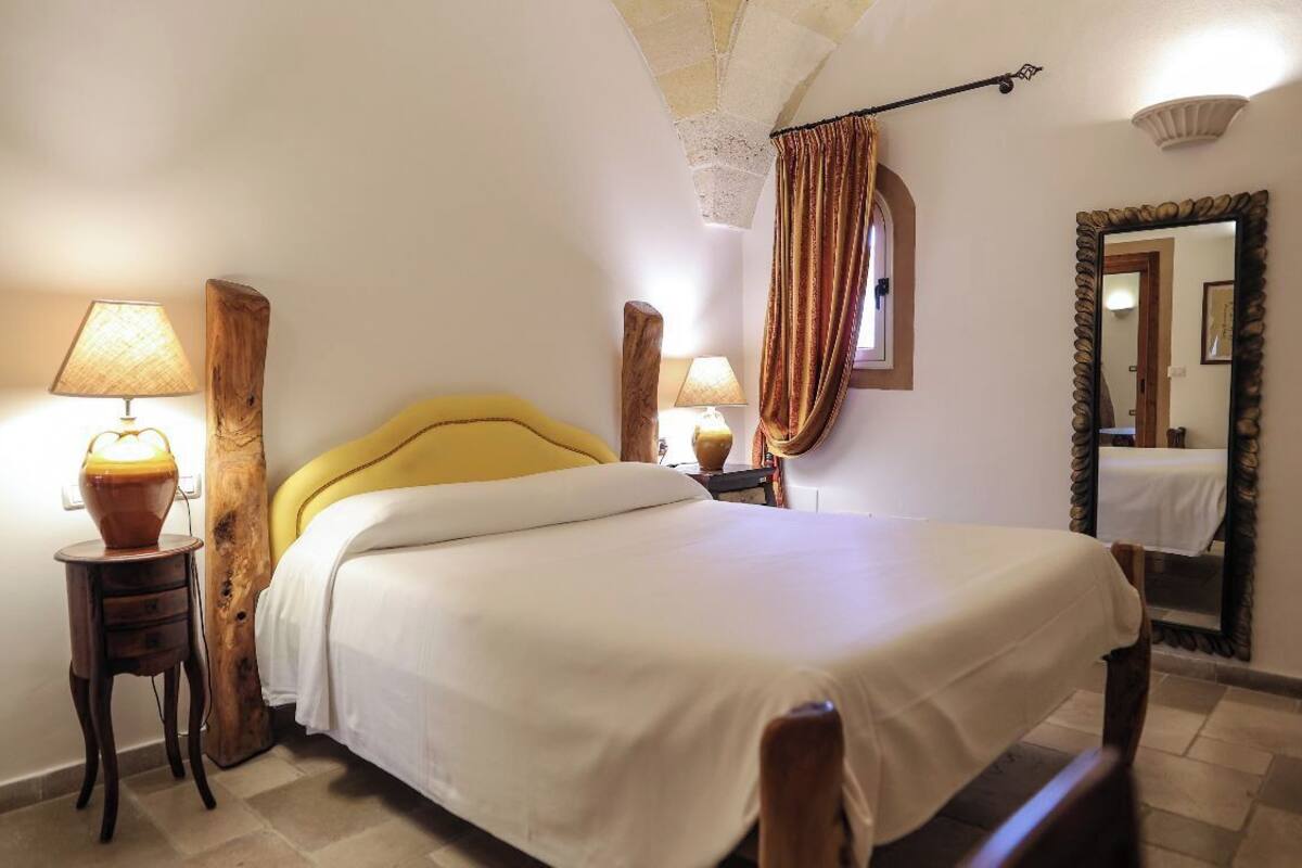 A neatly made bed with a yellow headboard, two bedside tables with lamps, a mirror with a decorative frame, and a window with curtains.