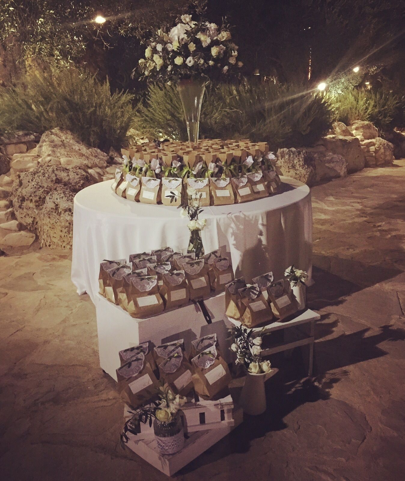 A dessert table with multiple tiers of cakes and small gift boxes, set on a sandy outdoor area with rocks and plants in the background.