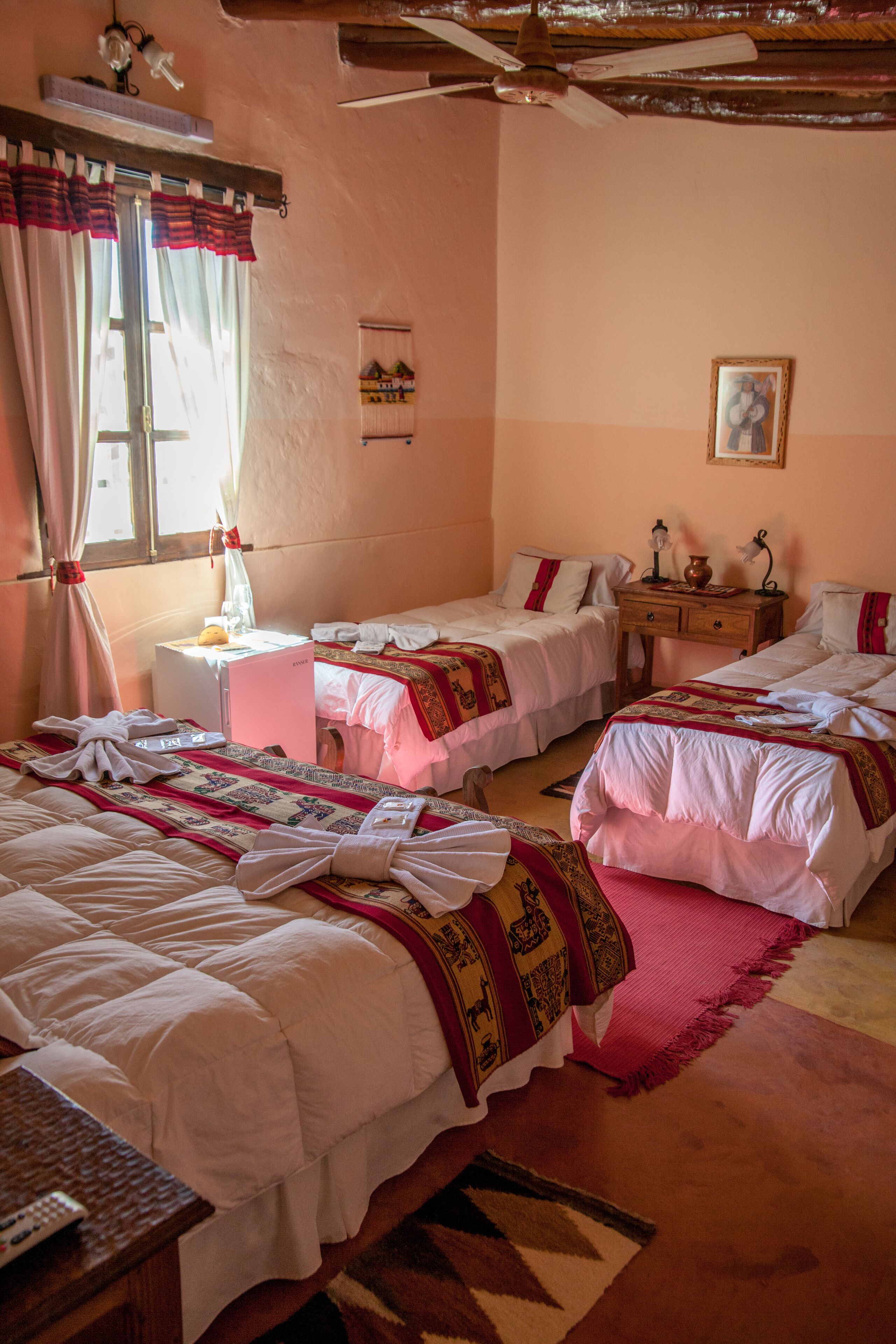 Quarto de hotel com duas camas, uma pequena mesa, mesa de cabeceira, janela com cortinas e ventoinha no teto.