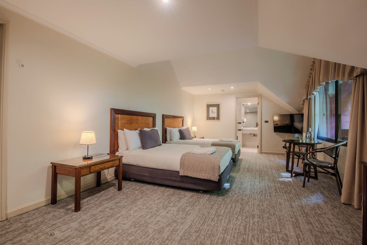 Une chambre d’hôtel avec deux lits, un bureau, une chaise, une télévision et une salle de bain visible à travers une porte ouverte.