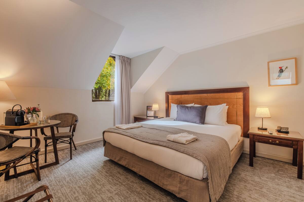 Une chambre d’hôtel avec un grand lit, un bureau avec une chaise, une petite table, une table de chevet et une fenêtre avec des rideaux.