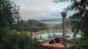 Un jacuzzi de madera con un ventil de vapor, rodeado de follaje y vistas a un lago.