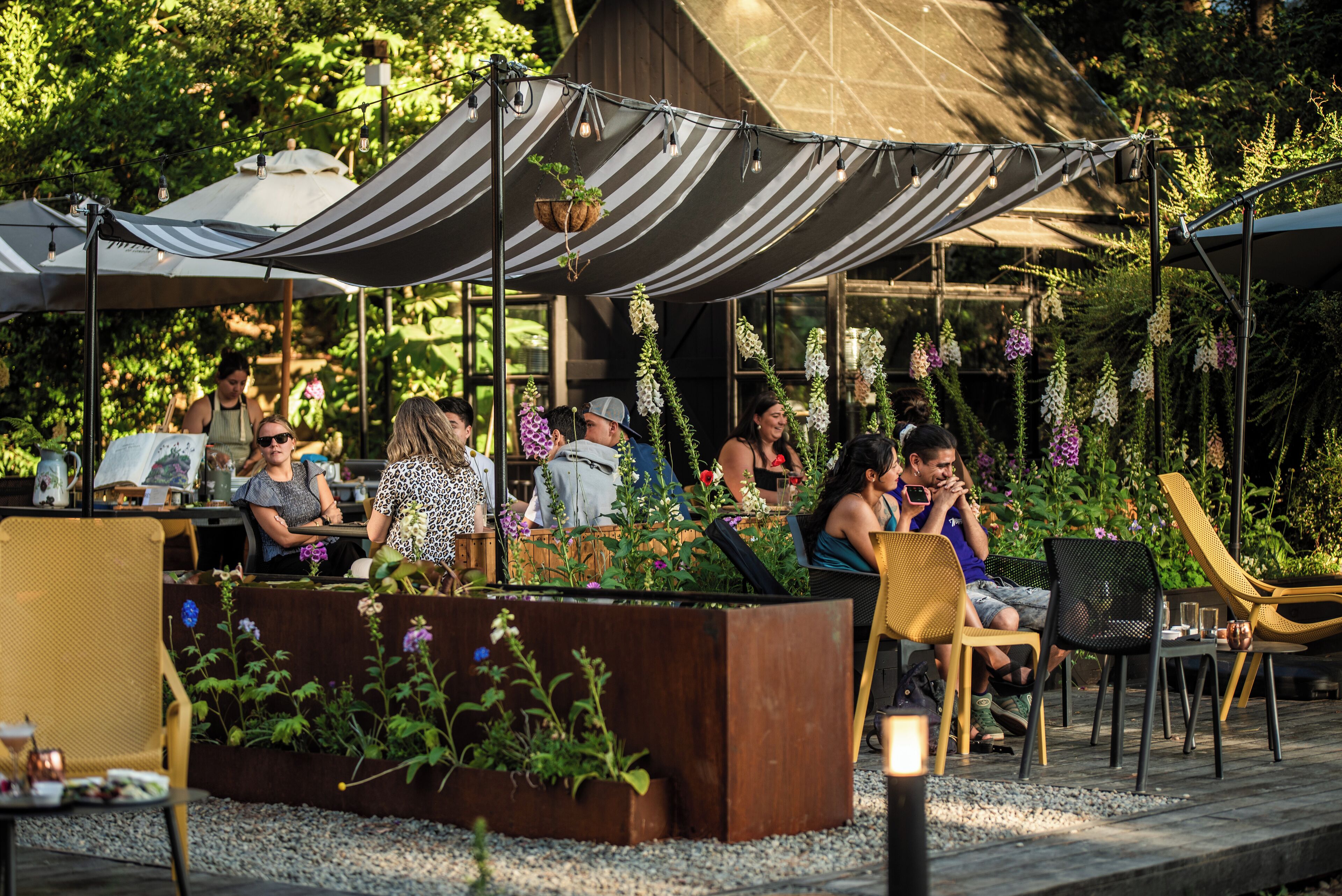 Een terras met tafels en stoelen onder gestreepte luifels, omgeven door groen en bloeiende planten.