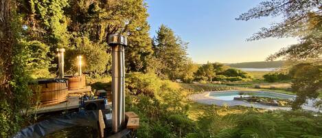 A hot tub with a wooden deck and a tall metal chimney, surrounded by lush greenery and a clear view of a lake and hills in the distance.