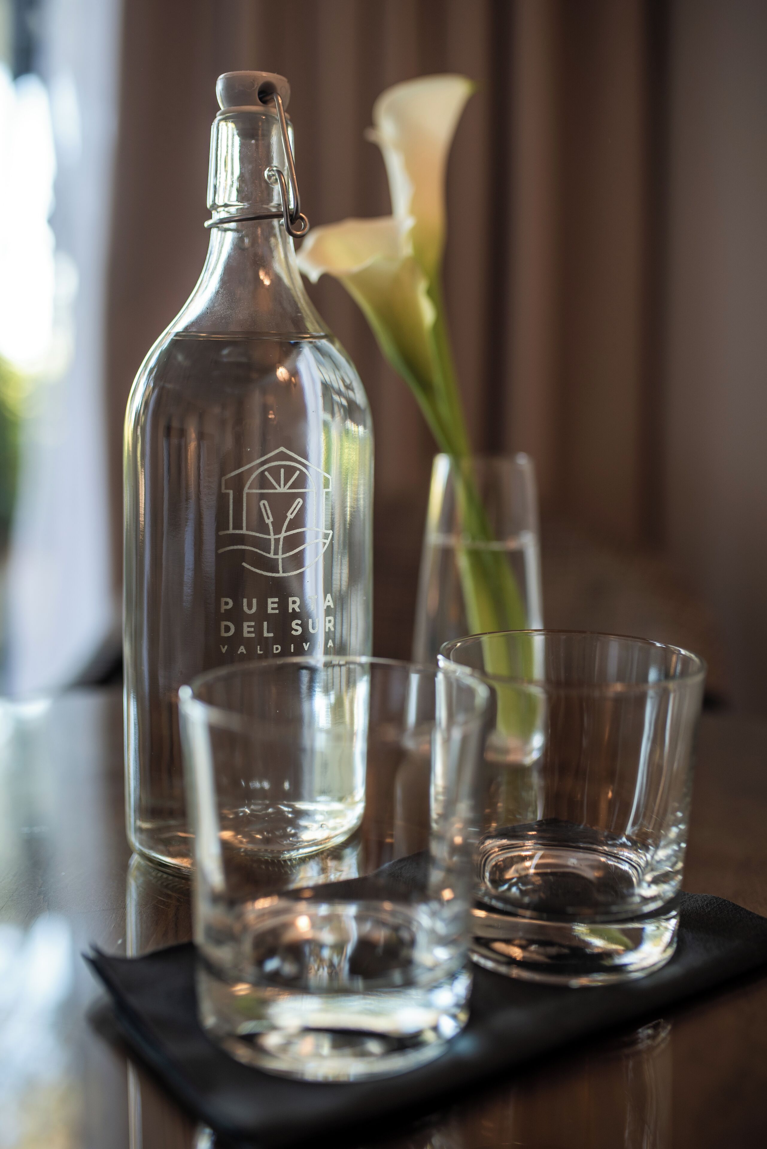 A clear bottle with embossed text, two clear glasses, and a white flower in the background.
