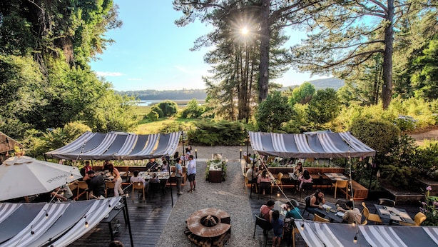 Un área de comedor al aire libre con mesas y sillas bajo toldos a rayas, una chimenea central y vistas a un lago y colinas boscosas.