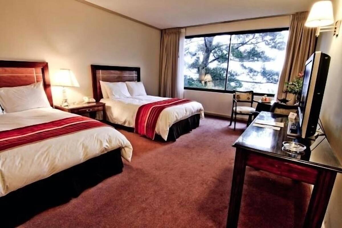 Une chambre d’hôtel avec deux lits, un bureau, une chaise et une grande fenêtre donnant sur la forêt.