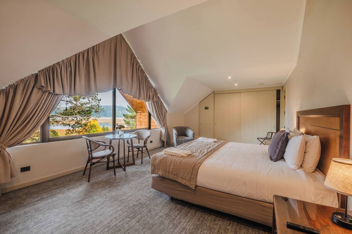 Une chambre d’hôtel avec un grand lit, un bureau, deux chaises, une vue sur l’extérieur et une tête de lit en bois.