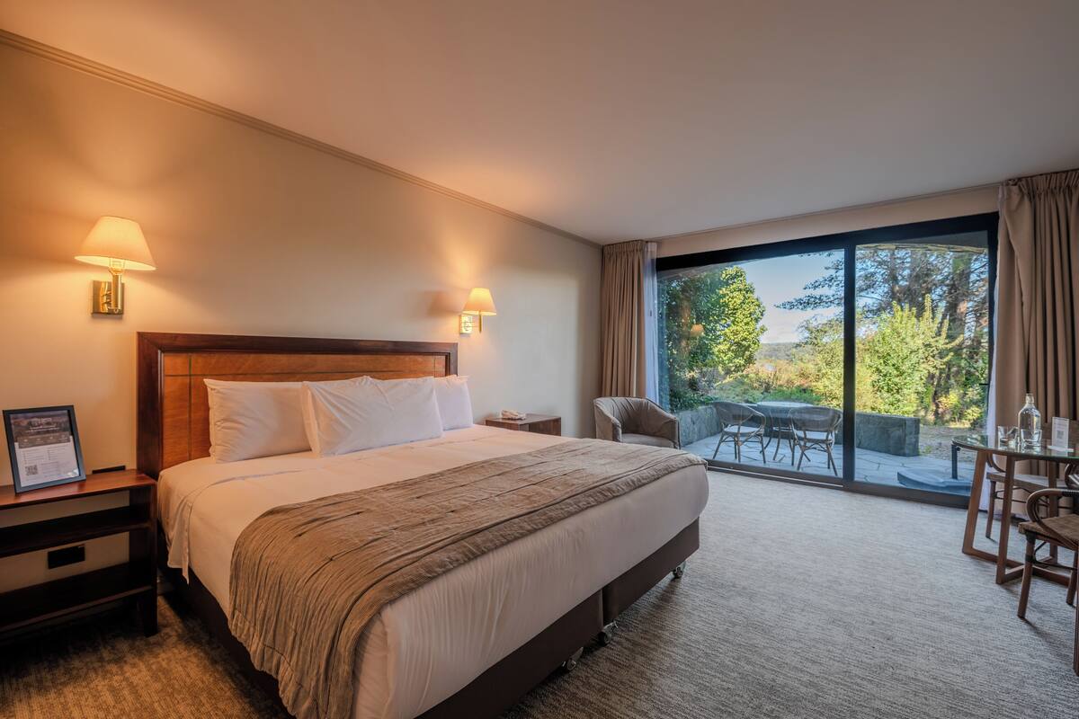 Une chambre d’hôtel avec un grand lit, deux lampes de chevet, une table de chevet, un bureau avec une chaise et une vue sur l’extérieur grâce à une grande fenêtre.