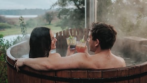 Dos personas en un jacuzzi con vista a un lago y árboles.