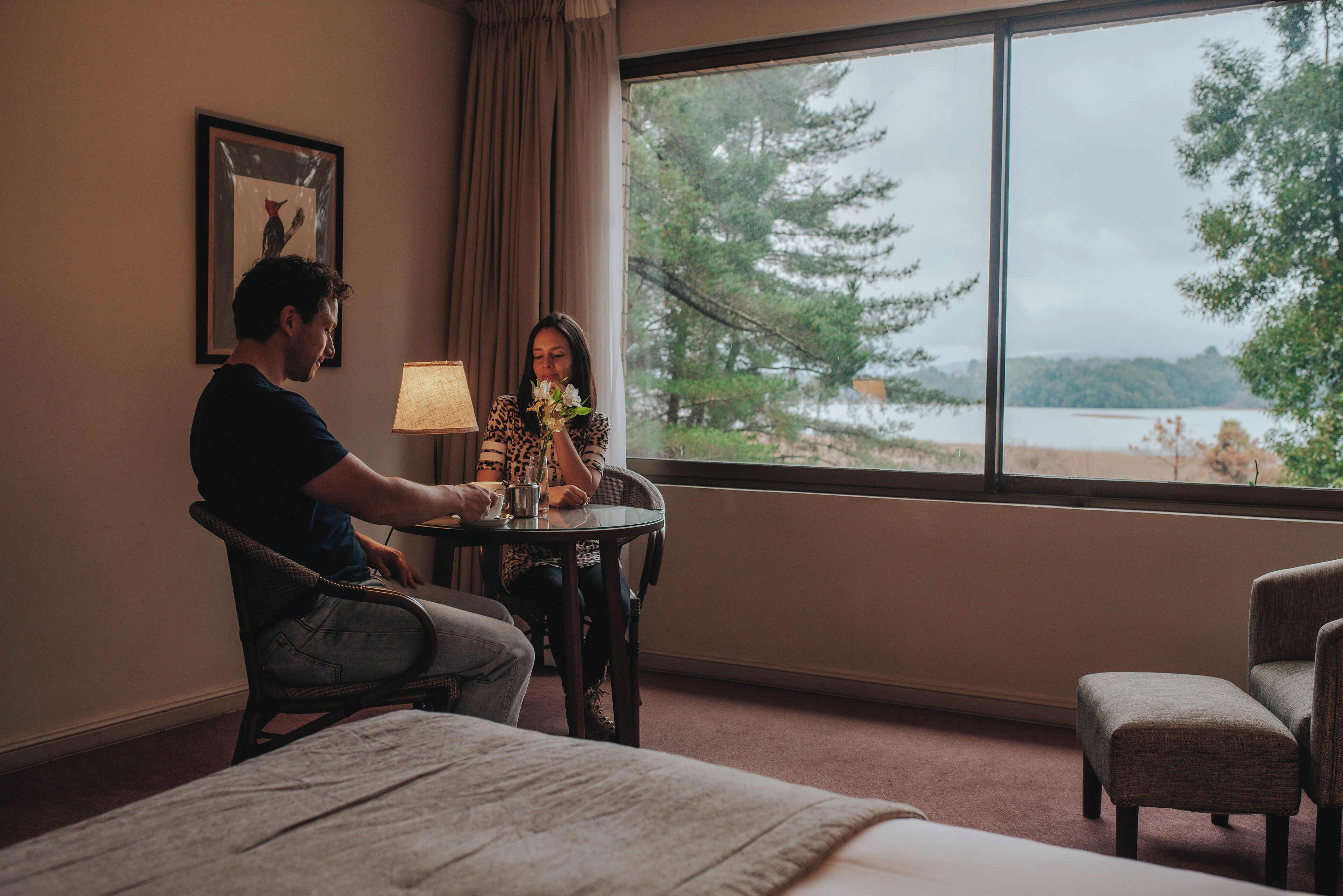 Habitación de hotel con un ventanal que ofrece vistas a árboles y una masa de agua. Hay una mesa con una lámpara y un jarrón de flores.