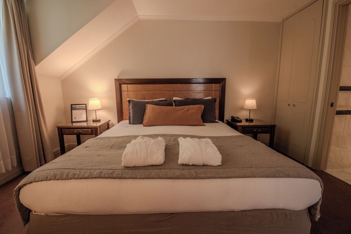 A hotel room with a large bed, two bedside tables with lamps, a wooden headboard, and a view of a door.