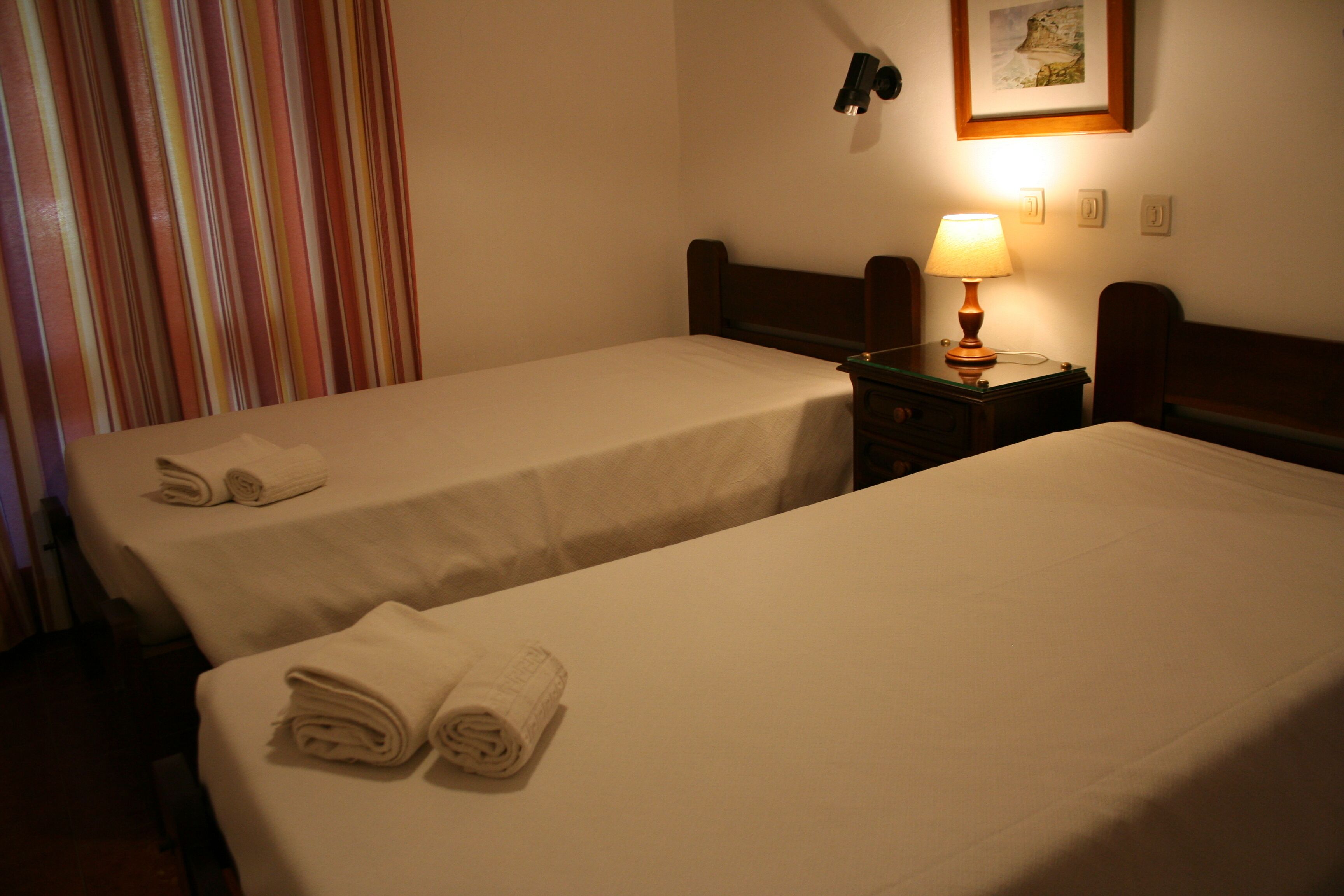 A hotel room with two beds, each with a neatly folded towel. There is a nightstand with a lamp between the beds.