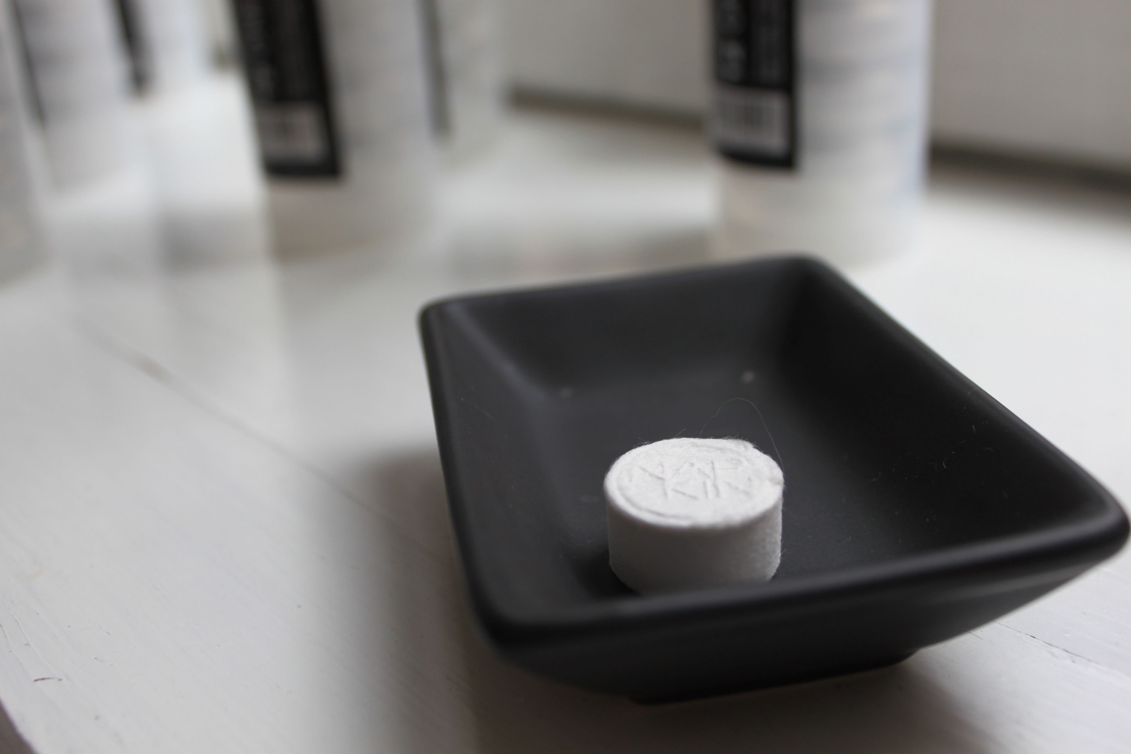 A small black square bowl containing a white pill on a white surface.