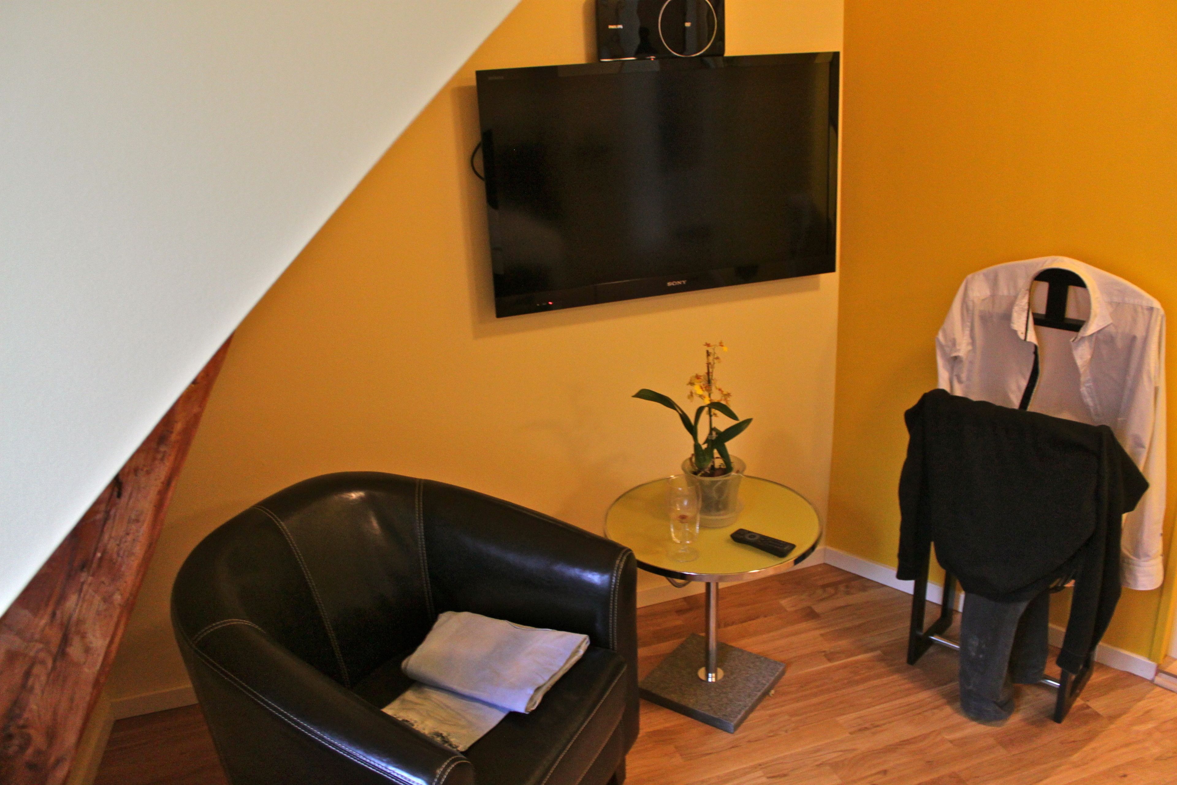 A room with a black leather sofa, a small round table with a vase, a flat-screen TV mounted on the wall, and a mannequin dressed in a white shirt and black pants.