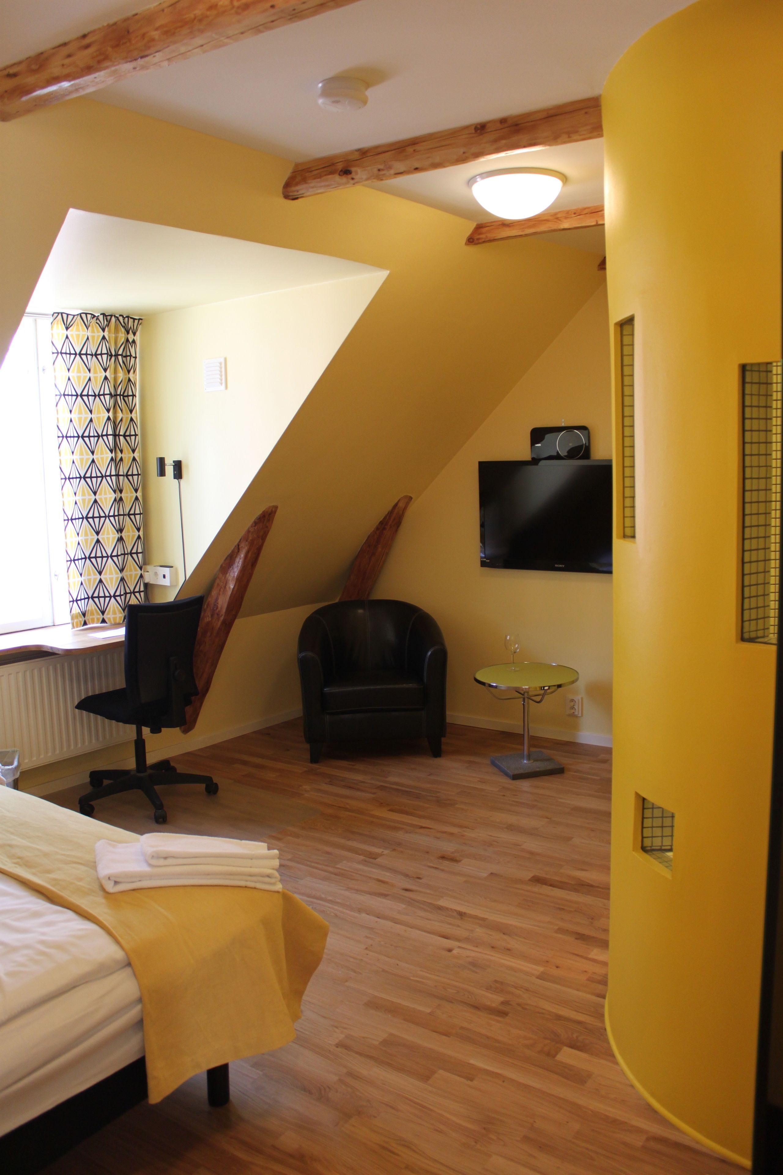 A room with a bed, a chair, a small table, a TV mounted on the wall, and a window with patterned curtains.