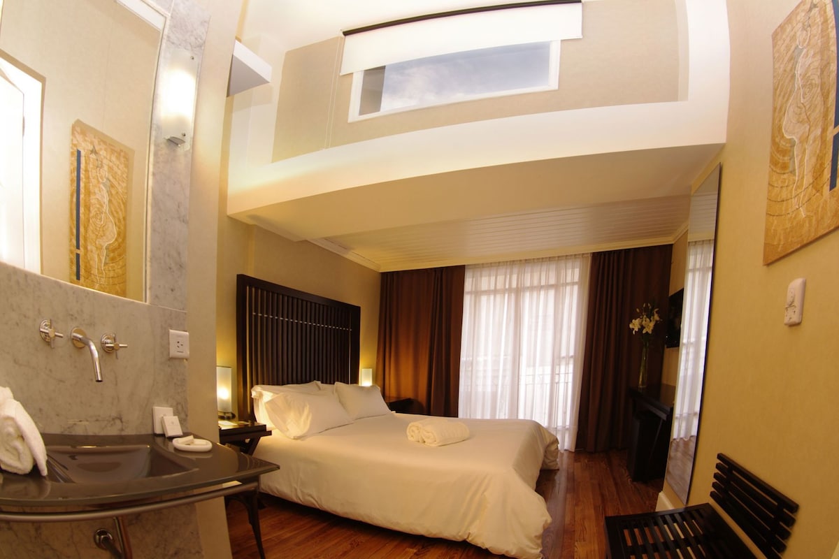 A hotel room with a bed, a bathroom with a sink, and a large window.