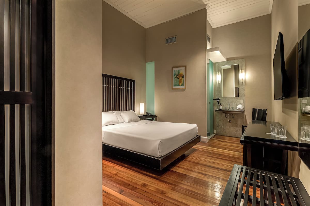 A hotel room with a bed, bedside table, and a desk with a chair. There is a mirror above the desk and a window with blinds.