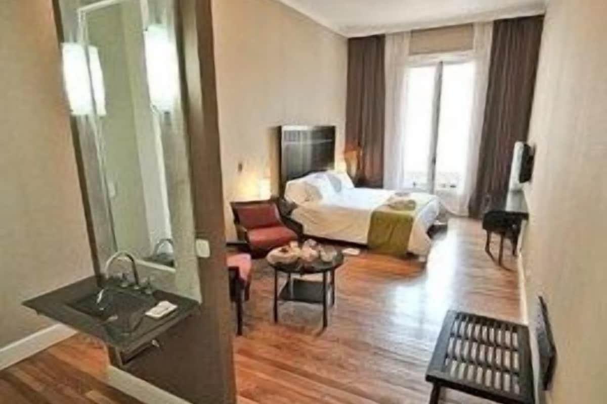 A hotel room with a wooden floor, a bed, a desk, a chair, a mirror, and a window with curtains.