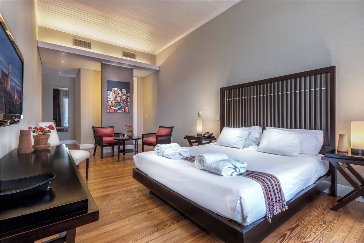 A hotel room with a large bed, a desk, a chair, a small table, and a painting on the wall.