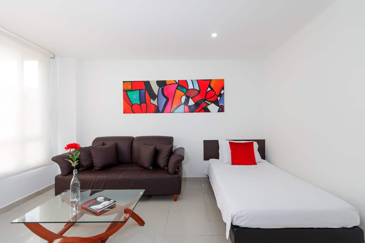 Een moderne hotelkamer met een glazen salontafel, een bruine leren bank, een wit bed met rode kussens, en een kleurrijk abstract schilderij aan de muur.