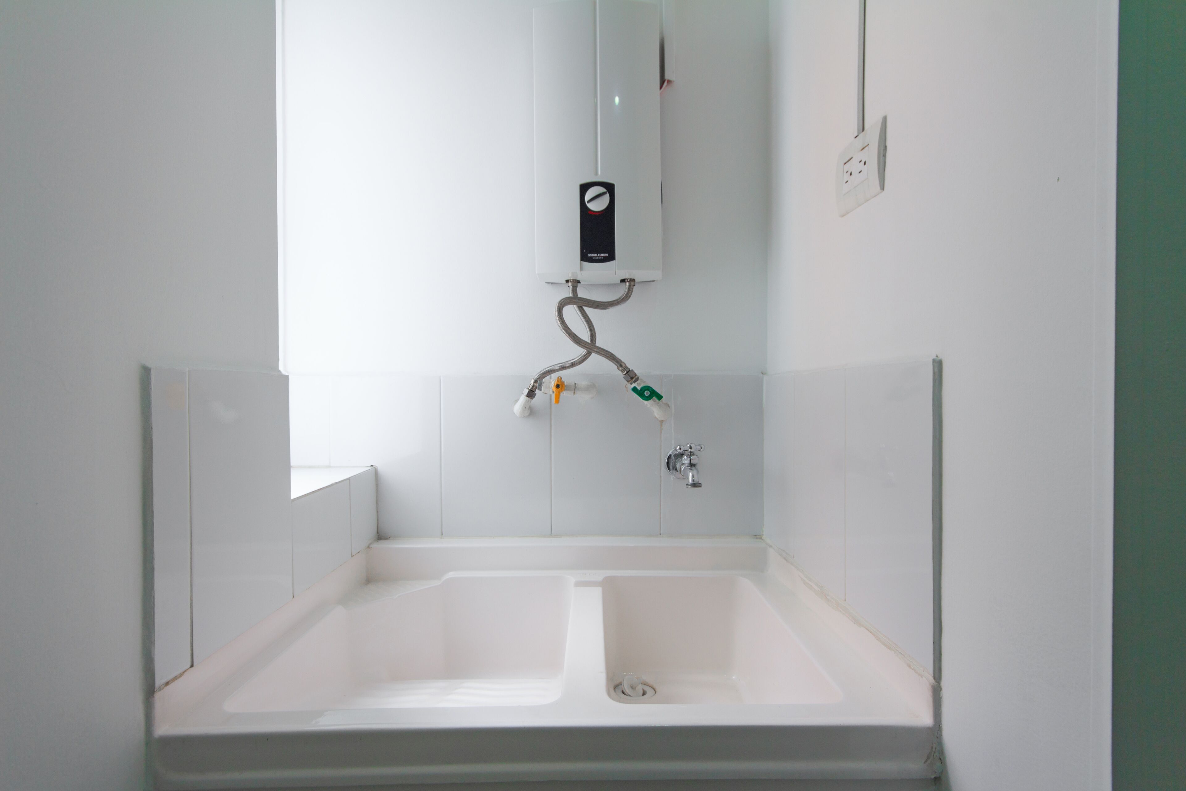 Un baño con una bañera blanca, un inodoro blanco y un lavamanos blanco. Cuenta con un calentador blanco instalado en la pared y un enchufe eléctrico blanco.