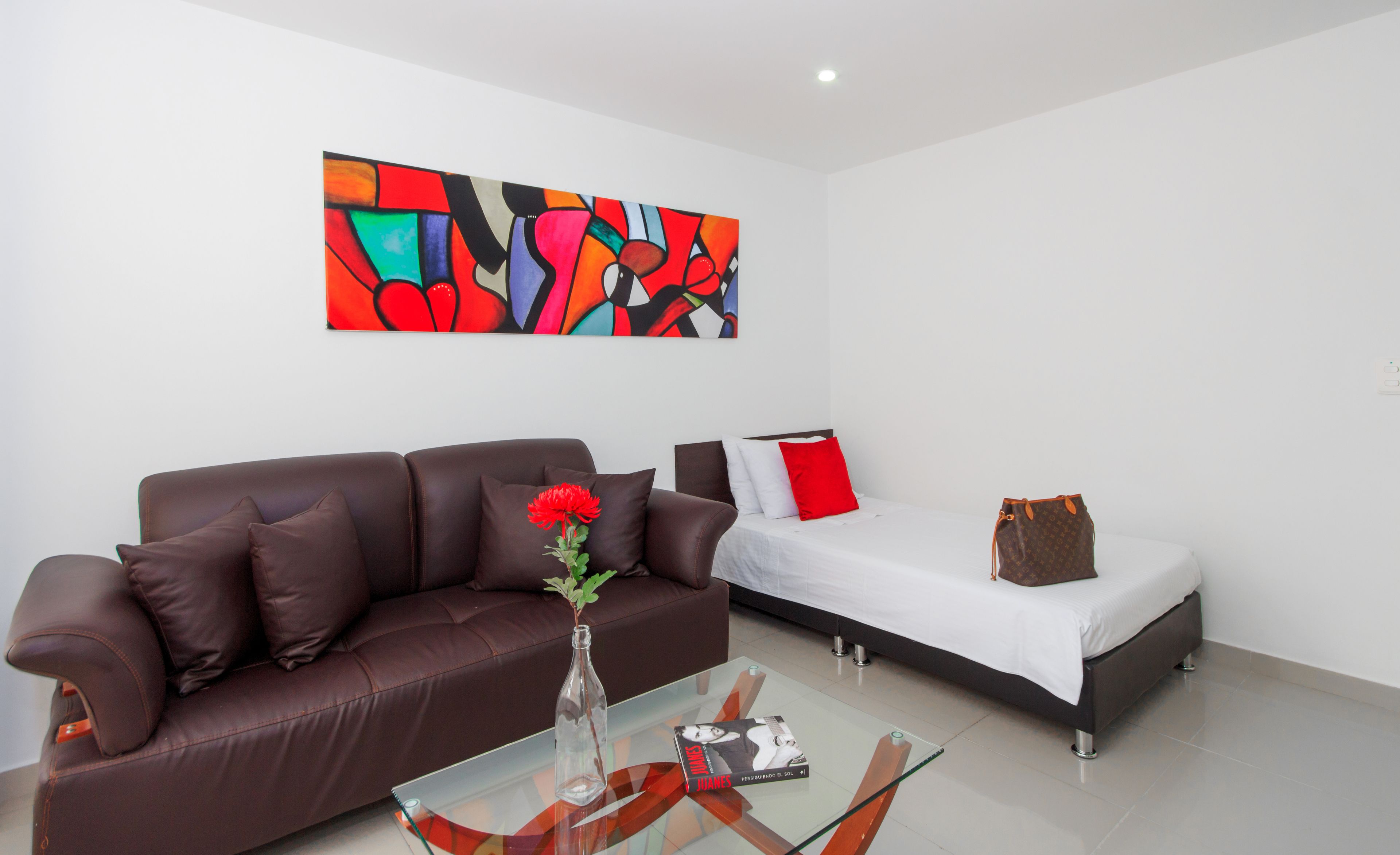 A modern living room with a brown leather sofa, a glass coffee table, and a bed with white bedding and red pillows. A colorful abstract painting adorns the wall above the sofa.