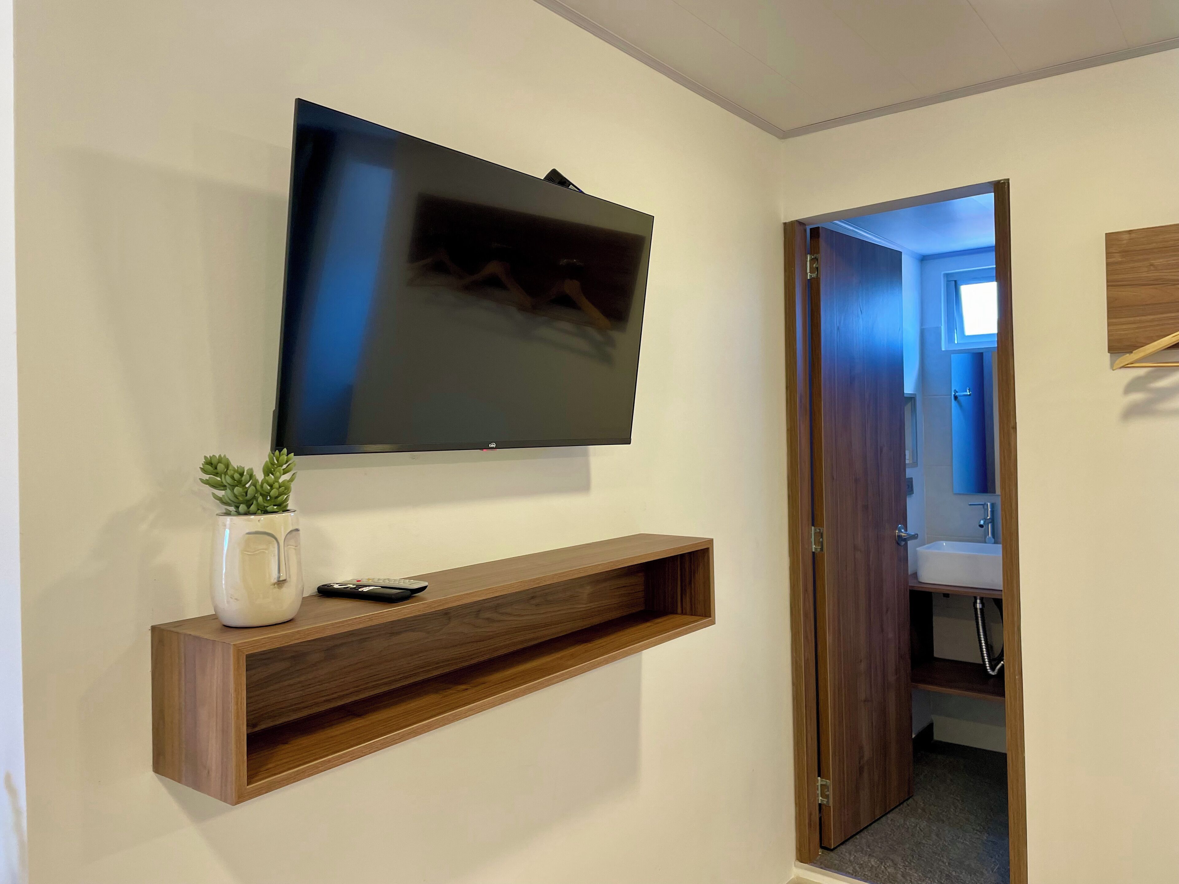 Un téléviseur fixé au mur, au-dessus d’une étagère en bois, avec une télécommande et une plante en pot. On aperçoit une salle de bain à travers une porte ouverte.