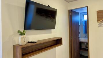 A wall-mounted TV above a wooden shelf with a remote control and a potted plant. A bathroom is visible through an open door.