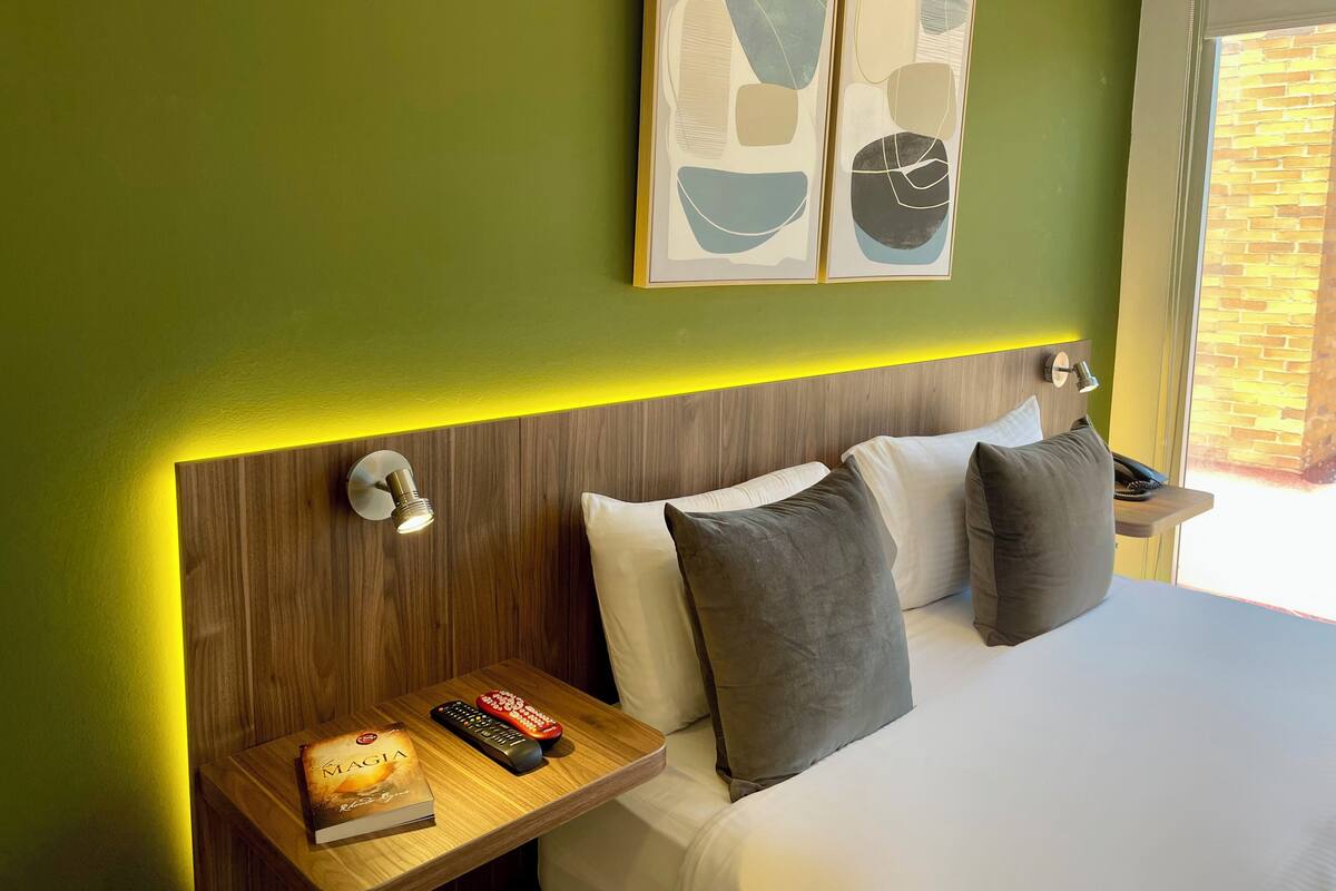 Een hotelkamer met een houten hoofdeinde, een nachtkastje met een afstandsbediening, een boek en een kussen.