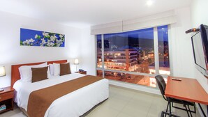 Une chambre d’hôtel avec un grand lit, un bureau et une vue sur le paysage urbain illuminé la nuit.