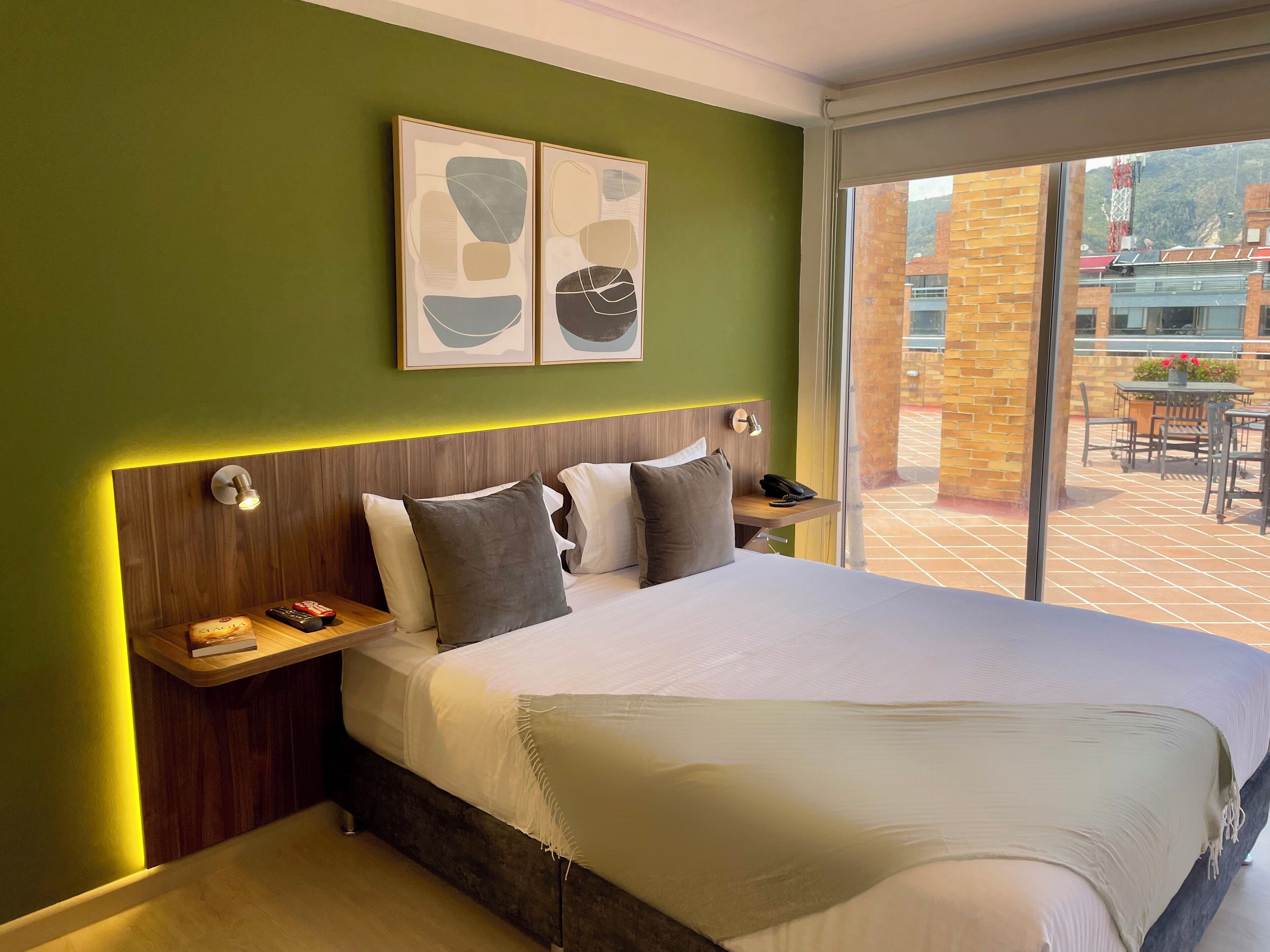 A modern hotel room with a large bed, a wooden headboard, a bedside table with a lamp, and artwork on the wall.