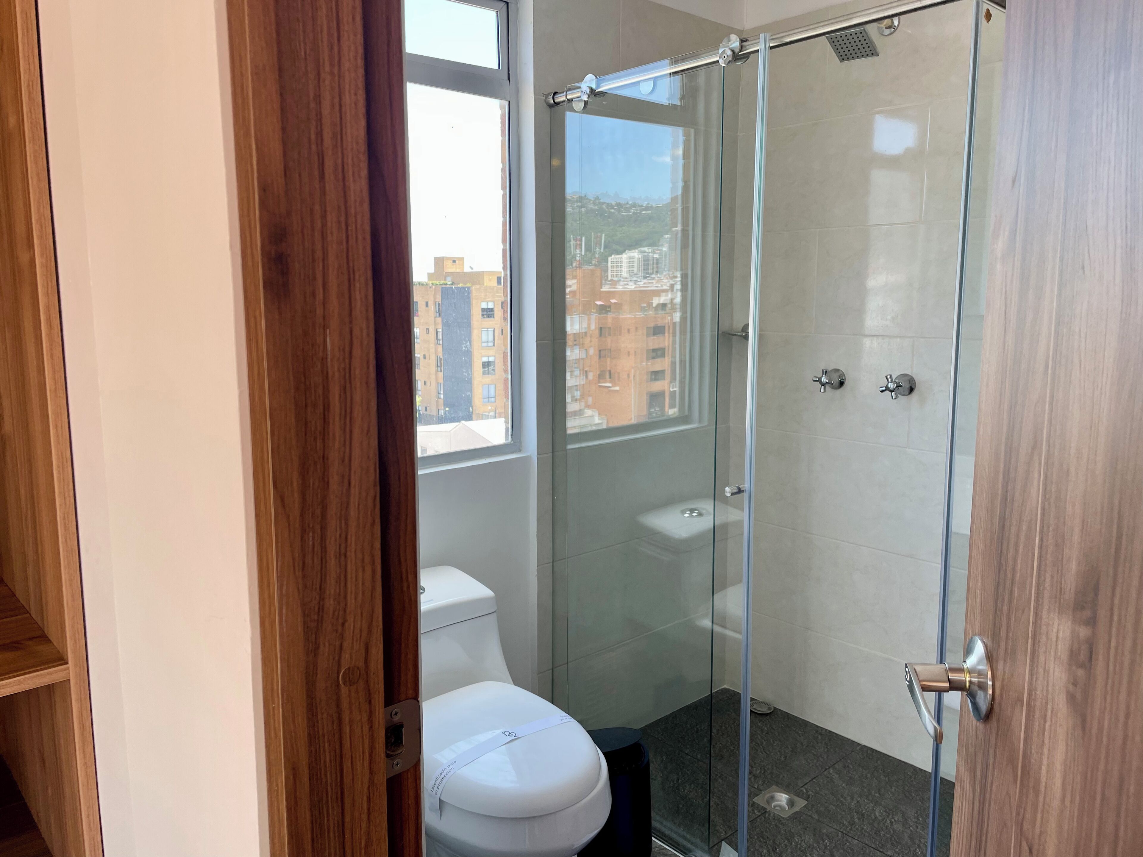 Un baño moderno con cabina de ducha de vidrio, inodoro blanco y vistas a edificios.