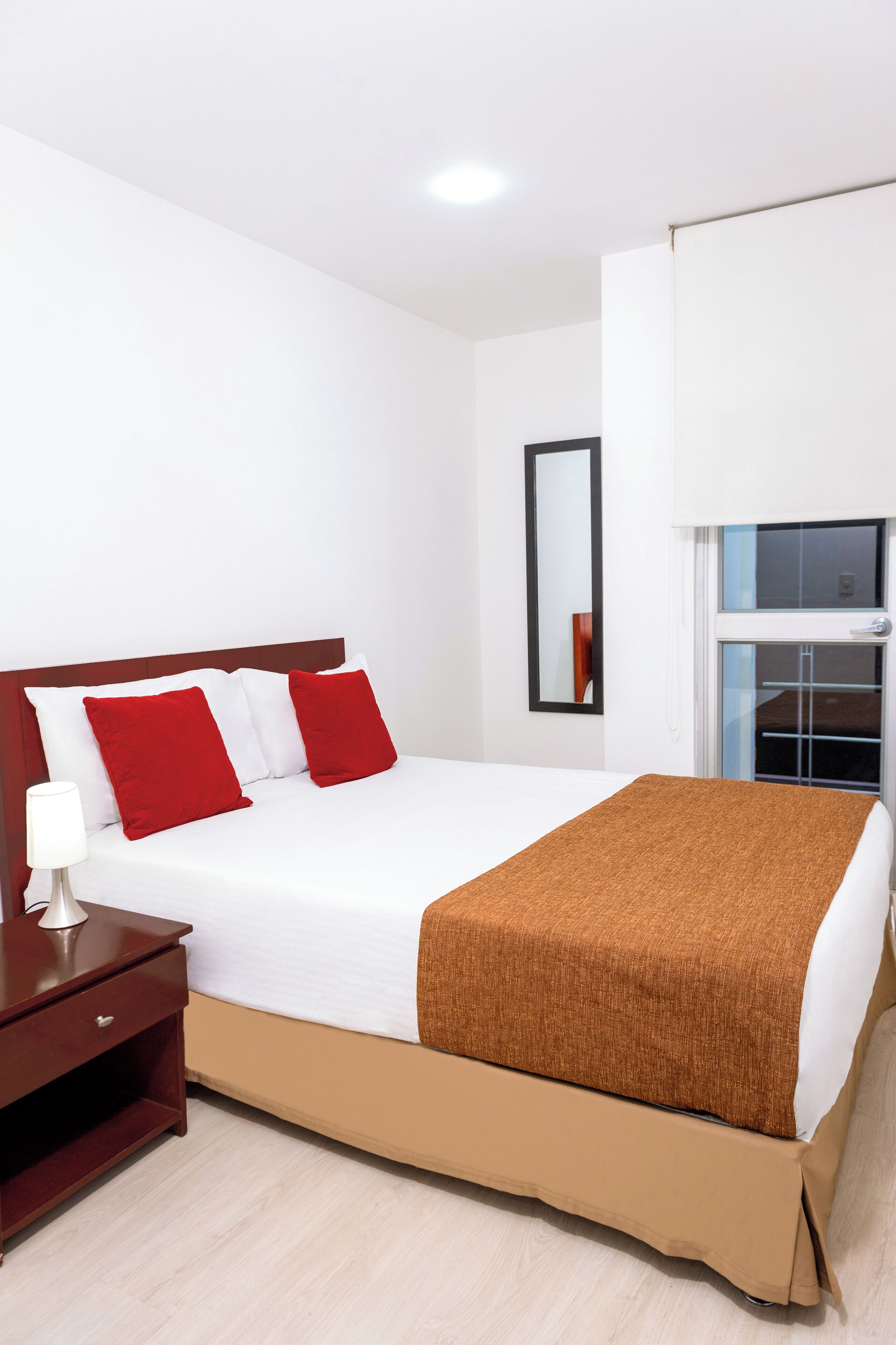 A neatly made bed with red and white pillows, a brown blanket, and a wooden nightstand with a lamp.