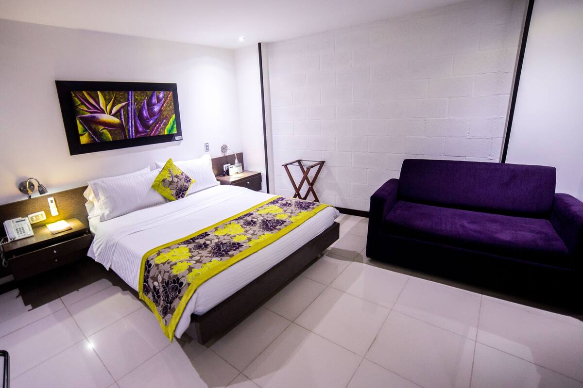 A modern hotel room with a bed, a purple sofa, a nightstand with a phone, and a painting on the wall.