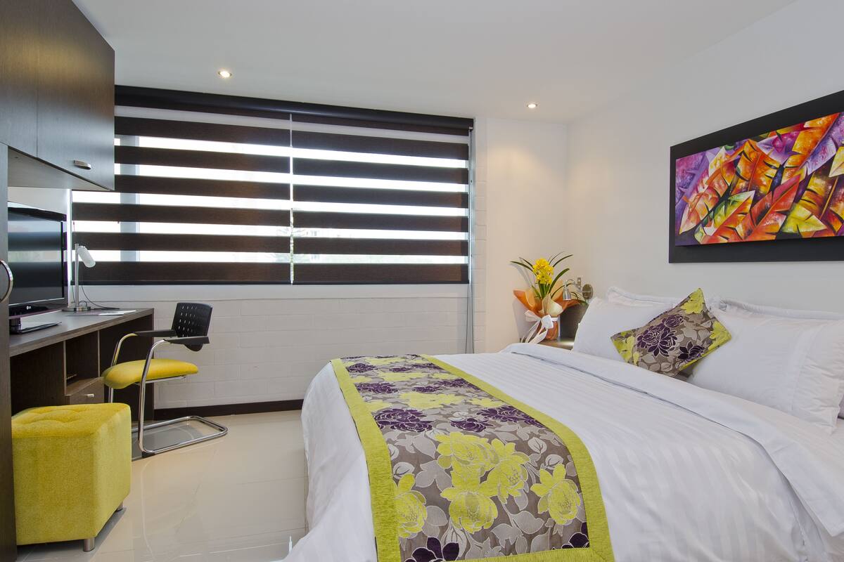 A modern hotel room with a large bed, a desk with a chair, and a window with blinds.