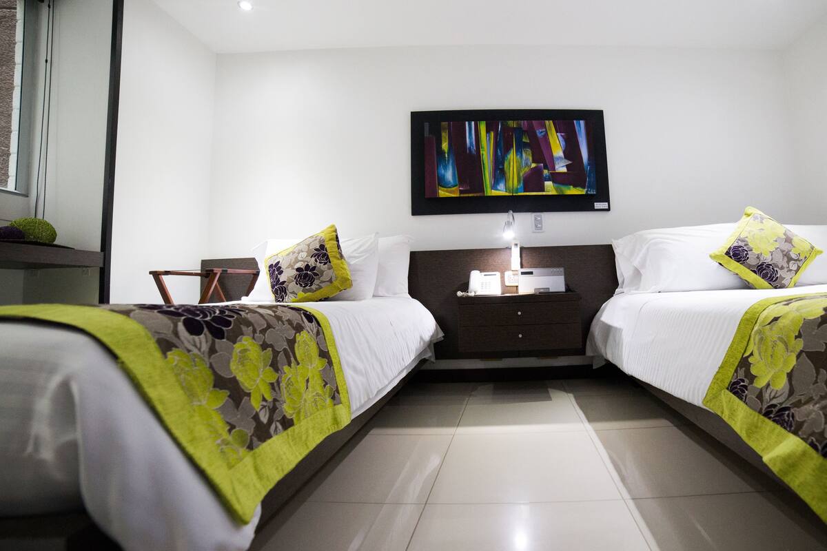 A hotel room with two beds, a nightstand, a flat-screen TV, and a small table with a lamp.
