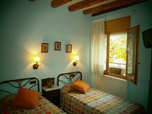 A room with two single beds, each with an orange pillow, a small bedside table, and a lamp. There are three framed pictures on the wall, and a window with a curtain.