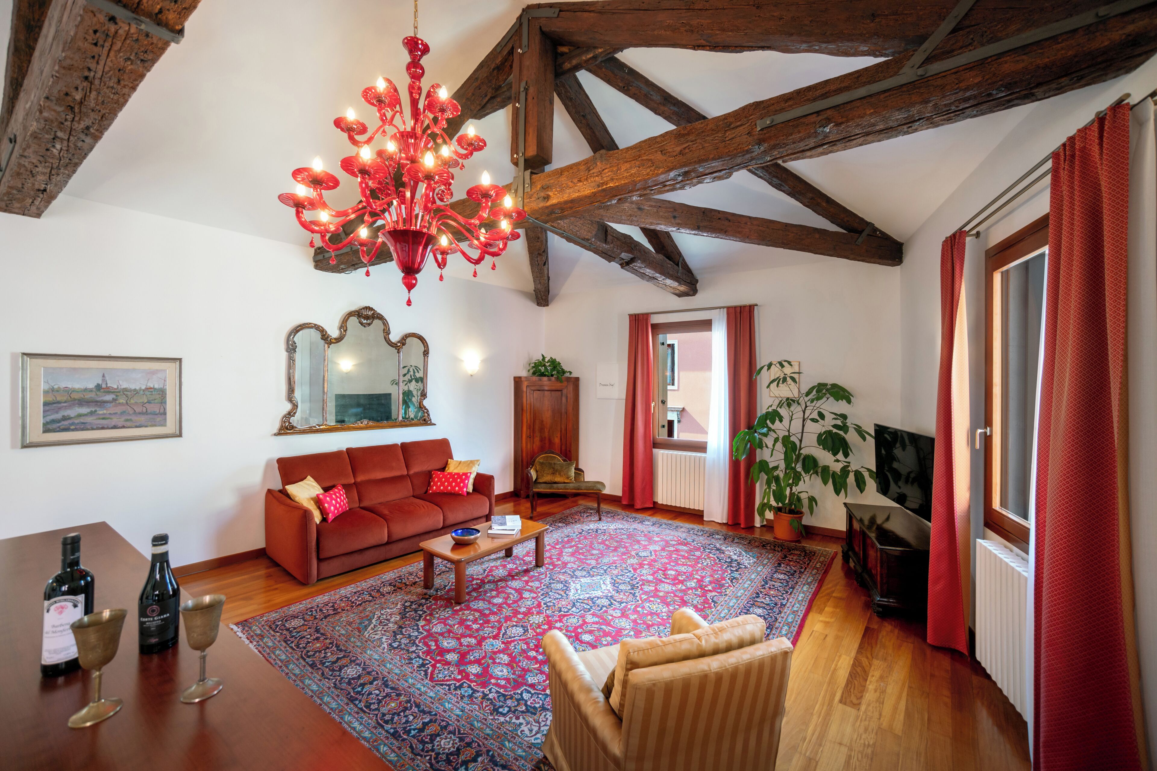 Un salon chaleureux avec un canapé rouge, des poutres en bois, un lustre, une grande moquette et un téléviseur.