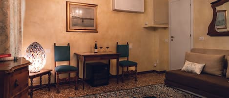 Ein gemütliches Wohnzimmer mit einer Couch, zwei Sesseln, einem Tisch mit Flasche und Glas, einer Deckenleuchte, einer Klimaanlage und einem gemusterten Teppich.