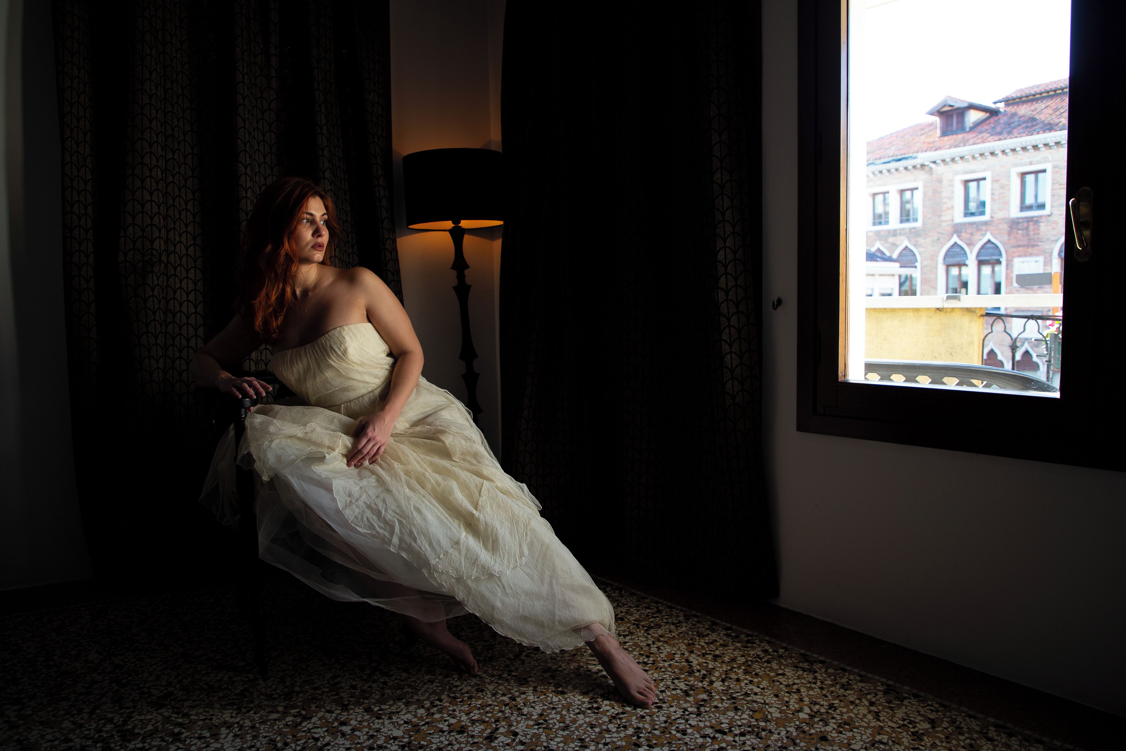 A woman in a white dress sitting on a chair in a dimly lit room with a window showing a building outside.