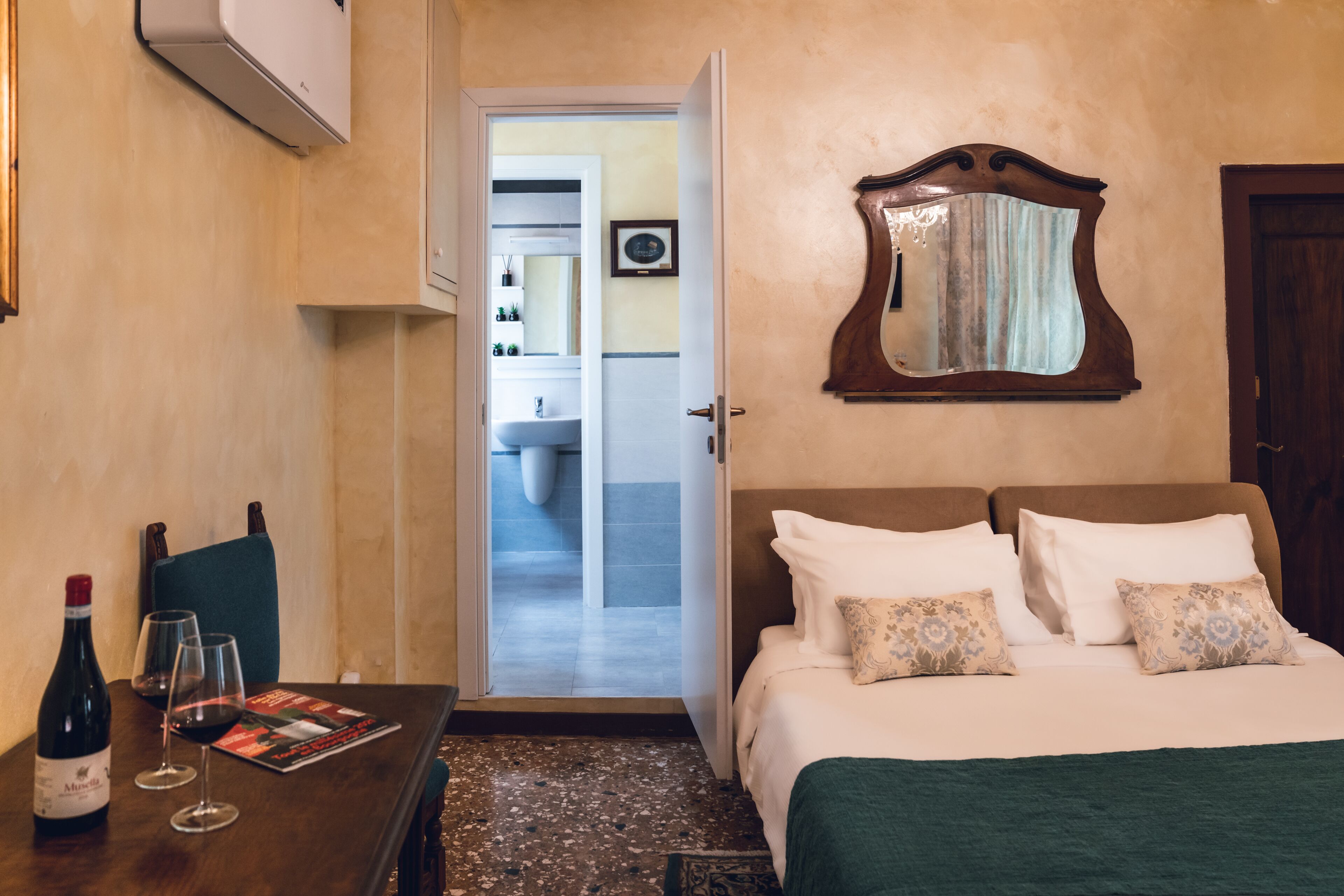 Une chambre d’hôtel avec un lit, une table sur laquelle se trouvent une bouteille de vin et des verres, un miroir et une vue sur la salle de bain.