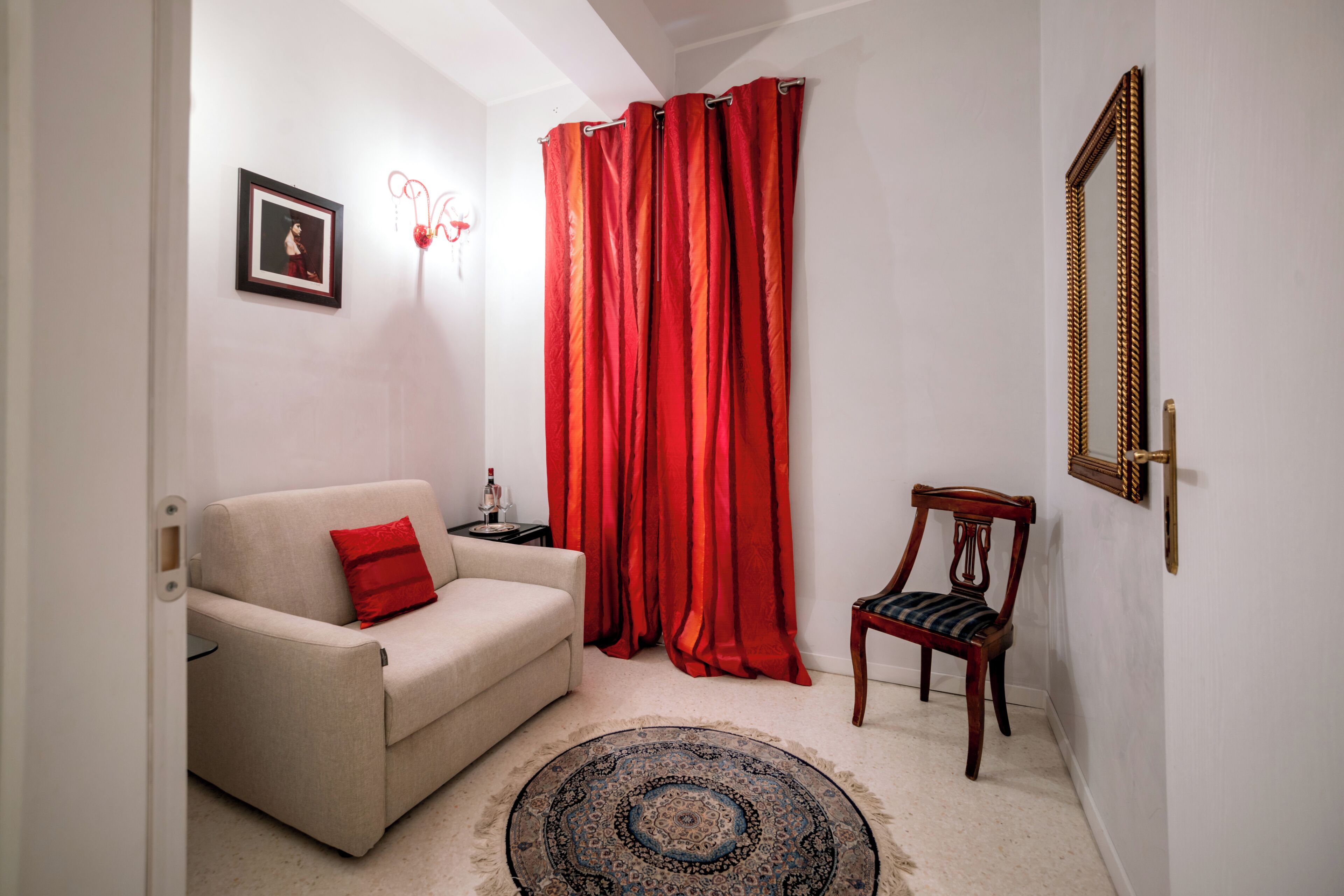 A living room with a beige sofa, a red cushion, a patterned rug, a wooden chair, a red curtain, a framed mirror, and a small table with a lamp.