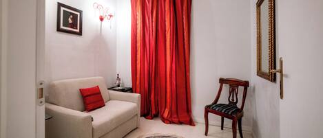 A living room with a beige sofa, a red cushion, a patterned rug, a wooden chair, a red curtain, a framed mirror, and a small table with a lamp.