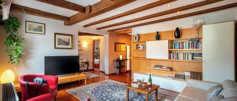 A living room with a TV, red chair, white sofa, wooden beams, bookshelf, and a patterned rug.