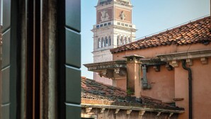 A view through a window of a building with a clock tower, a tiled roof, and a stone facade. - Palazzo San Luca (Venice)