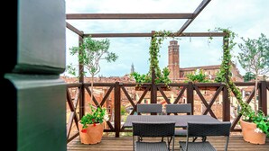 A wooden deck with two tables and chairs, surrounded by potted plants, overlooking a cityscape with historic buildings and a tower. - Palazzo San Luca (Venice)