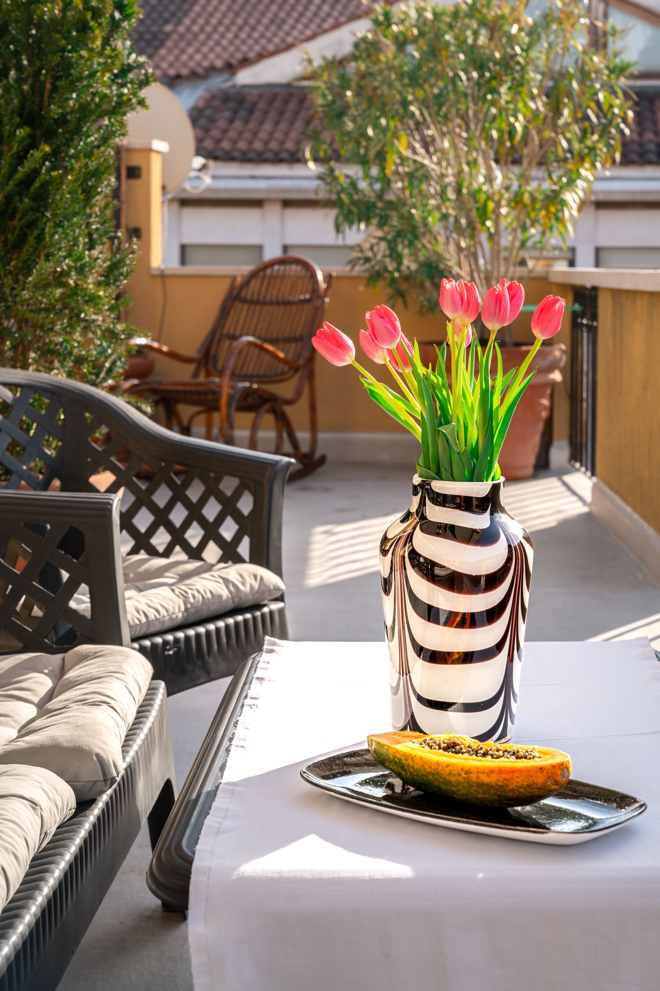 Une terrasse avec des meubles d’extérieur, un vase rayé contenant des tulipes roses et une assiette avec des fruits.