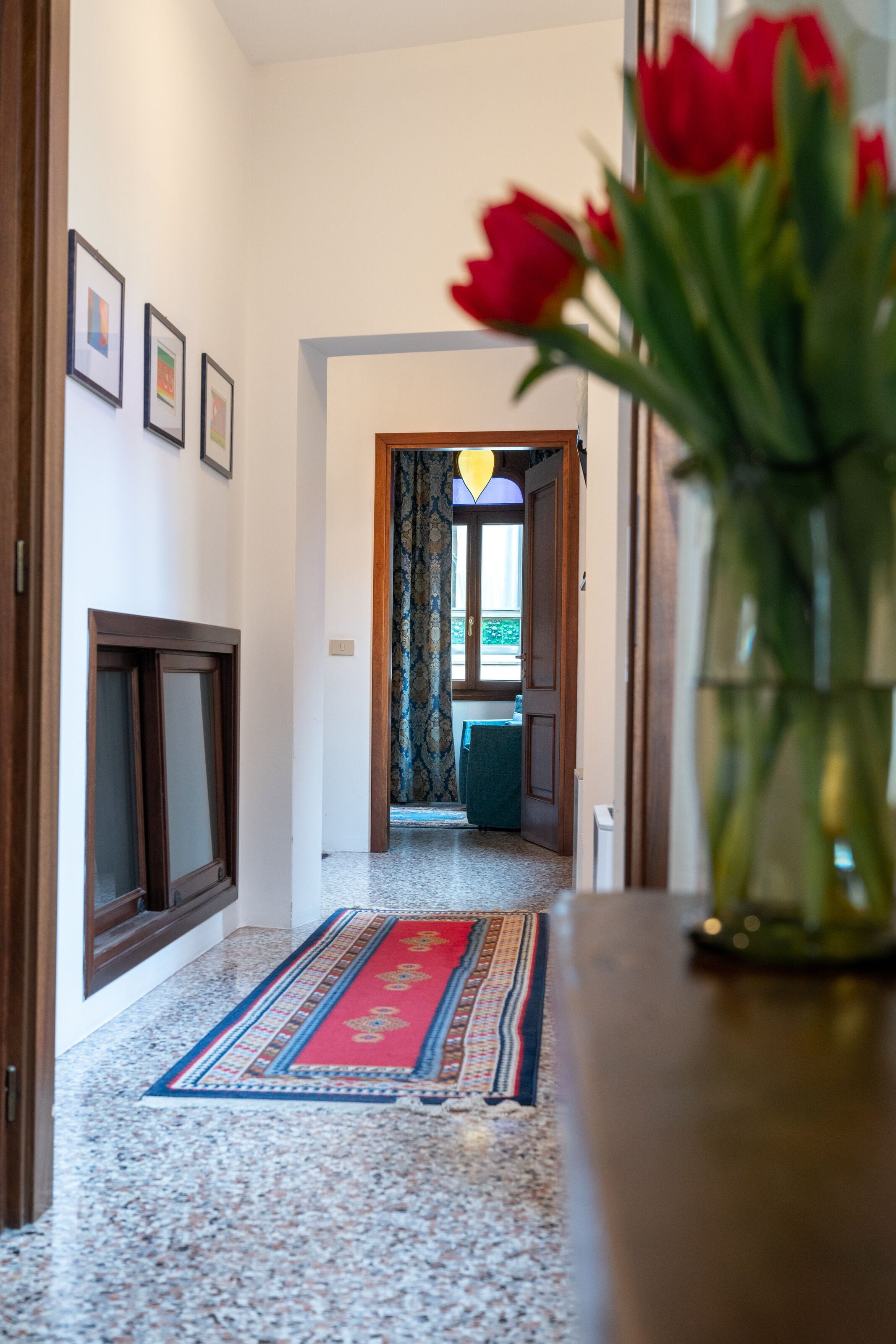 A hallway with a patterned rug, a wooden door, and a vase with red flowers.