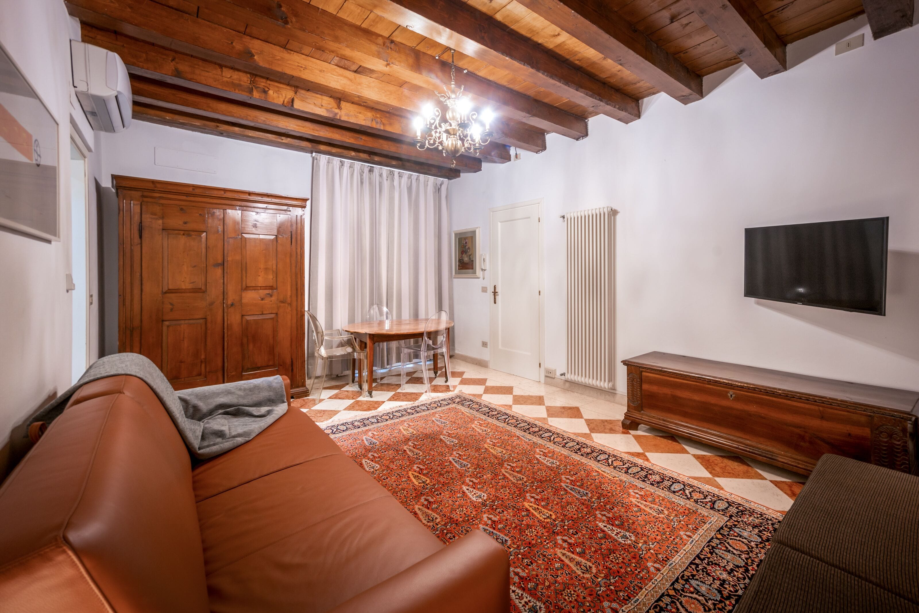 A living room with a brown leather sofa, a patterned rug, wooden beams on the ceiling, a television, and a wooden cabinet.